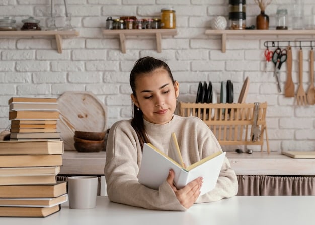 A student studying with books spread open on a desk, bathed in soft natural light. The student is smiling gently, with a laptop, coffee mug, and a pair of glasses nearby. The background is a blurred library scene with bookshelves and other students.
